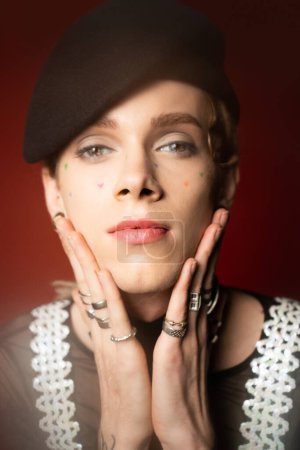 Foto de Portrait of nonbinary person in black beret and silver finger rings holding hands near face and looking at camera on dark red background - Imagen libre de derechos