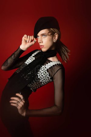 queer person in elegant attire touching black beret while posing with hand on hip on dark red background