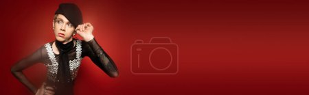 Photo for Young and stylish nonbinary person touching black beret and looking away on dark red background, banner - Royalty Free Image