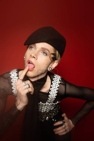 nonbinary person in stylish outfit touching tongue with finger while looking away on red background