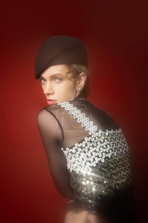 young nonbinary model in elegant top with sequins looking at camera on dark red background