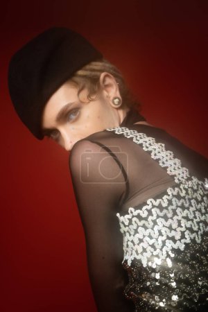 young nonbinary person in black beret and elegant top with sequins looking at camera on dark red background