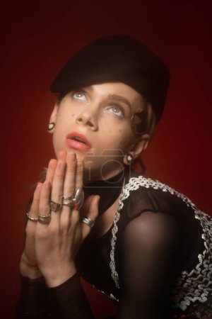 portrait of young nonbinary person in black beret and silver rings praying on dark red background