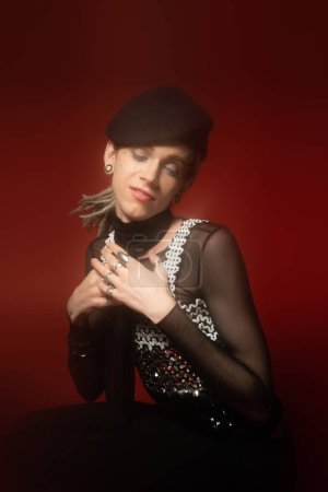 positive queer model in fashionable outfit and silver rings posing with closed eyes and hands near chest on dark red background