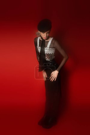 full length of blurred queer model in elegant attire standing with closed eyes on dark red background