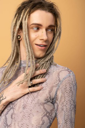 smiling queer person in top with snakeskin print touching dreadlock and looking away isolated on yellow