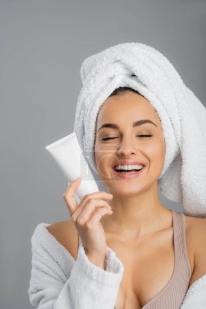 Smiling woman with towel on head holding cream isolated on grey 