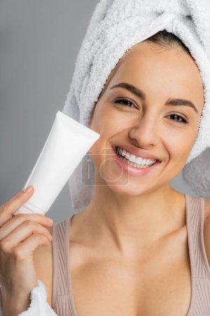 Smiling woman with towel on head holding cosmetic cream isolated on grey 