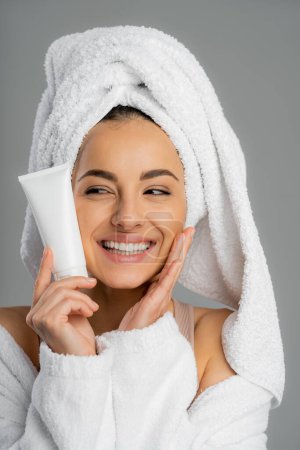 Positive young woman with towel on head holding cream and touching face isolated on grey 