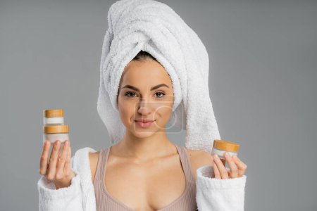 Young woman with towel on head holding creams isolated on grey 