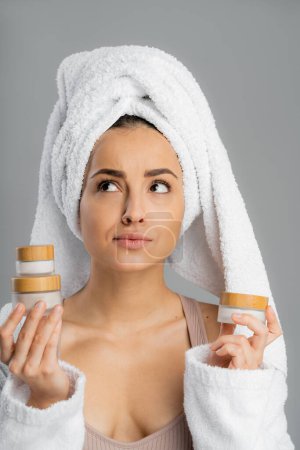 Pensive young woman with towel on head holding creams isolated on grey 