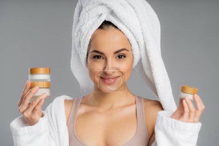 Cheerful young woman with towel on head holding cosmetic creams isolated on grey 
