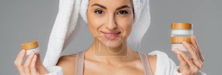 Smiling woman with towel on head holding cosmetic creams isolated on grey, banner 