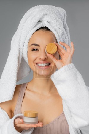 Positive woman with towel on head holding jars with cosmetic cream isolated on grey 