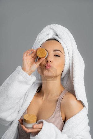 Foto de Woman in towel and bathrobe holding cosmetic cream and pouting lips isolated on grey - Imagen libre de derechos