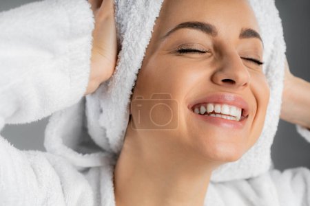 Cheerful young woman touching white towel on head isolated on grey 