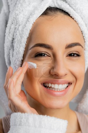 Smiling woman with towel on head applying face cream isolated on grey 