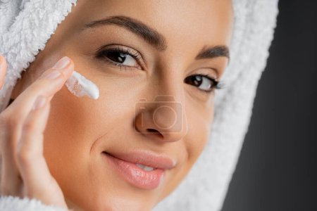 Close up view of smiling woman with towel on head applying face cream isolated on grey 