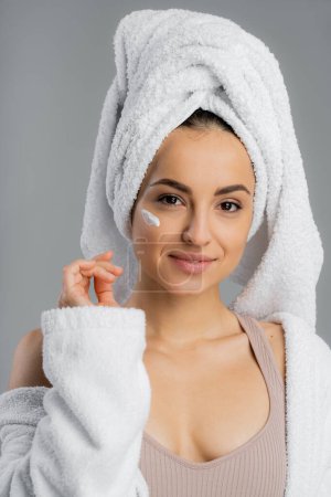 Pretty woman with towel on head and cream on face isolated on grey 