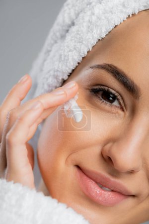 Cropped view of smiling woman with towel on head applying face cream isolated on grey 