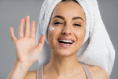 Cheerful woman with towel on head and cosmetic cream on cheek isolated on grey 