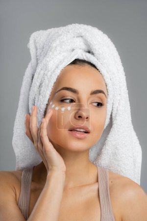Young woman with towel on head applying cream under eye isolated on grey 