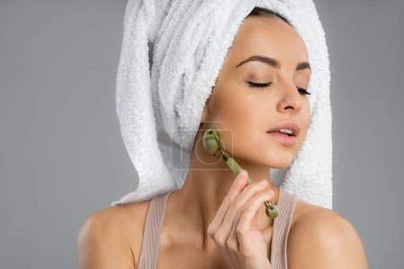 Woman with towel on head using jade roller isolated on grey 