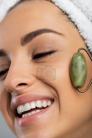 Close up view of smiling woman with towel on head massaging face with jade roller isolated on grey 