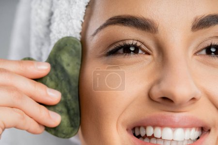 Close up view of cheerful woman with towel on head using gua sha isolated on grey 
