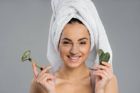 Smiling young woman with towel on head holding gua sha and jade roller isolated on grey 