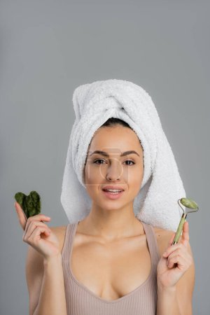 Young woman with towel on head holding jade roller and gua sha isolated on grey 
