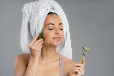 Pleased woman with towel on head using gua sha and holding jade roller isolated on grey 