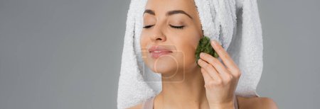 Pleased young woman with towel on head using gua sha isolated on grey, banner 