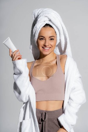 Smiling young woman in bathrobe and towel on head holding body lotion isolated on grey 