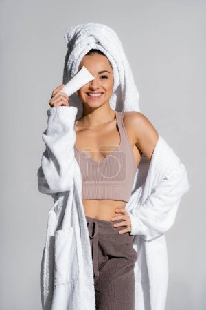 Cheerful woman in bathrobe holding body lotion near face isolated on grey 