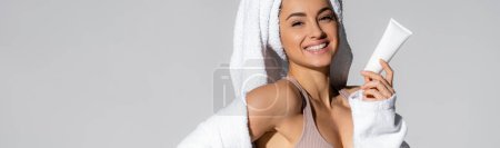 Cheerful young woman with towel on head holding cosmetic lotion isolated on grey, banner 