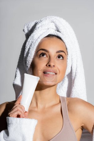 Pensive young woman with towel on head holding body lotion isolated on grey 