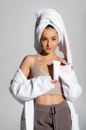 Young woman with towel on head holding cosmetic cream and looking at camera isolated on grey 