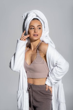 Young woman in bathrobe and towel on head touching face isolated on grey 