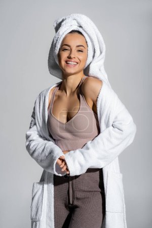Cheerful young woman in towel and bathrobe looking at camera isolated on grey 
