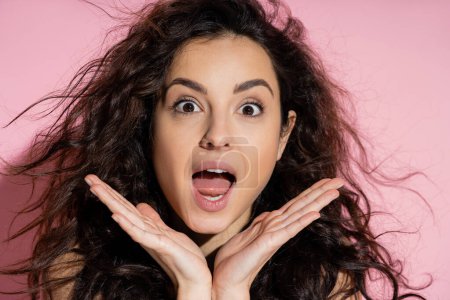 Téléchargez les photos : Portrait of shocked curly woman looking at camera on pink background - en image libre de droit