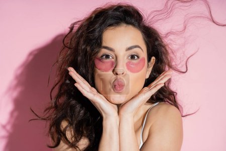 Curly woman with hydrogel eye patches pouting lips on pink background 
