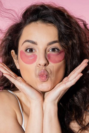 Portrait of curly woman with hydrogel eye patches pouting lips on pink background 