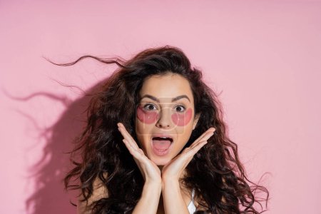 Excited brunette woman with eye patches on face looking at camera on pink background 