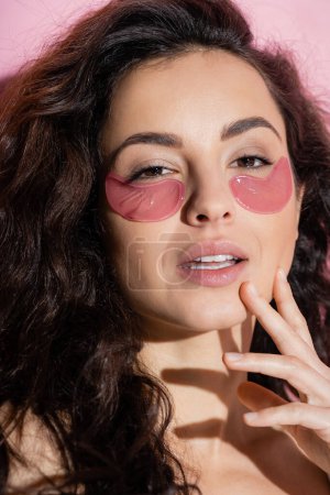 Portrait of curly woman with hydrogel eye patches looking at camera on pink background 