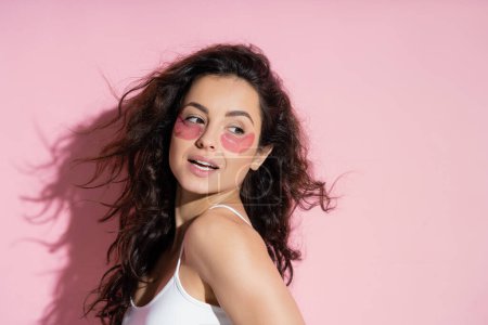 Curly young woman with hydrogel eye patches looking away on pink background 