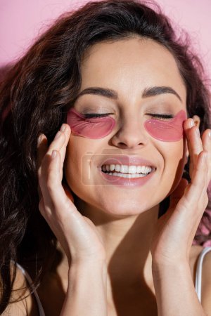 Curly woman touching hydrogel eye patches on face on pink background 