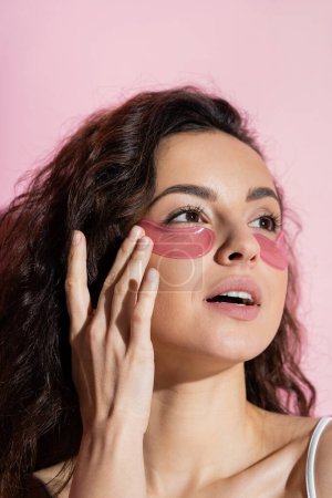 Young brunette woman applying hydrogel eye patch on face on pink background 