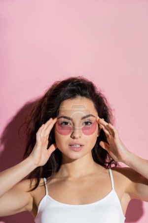 Foto de Pretty young woman in top touching hydrogel eye patches on pink background - Imagen libre de derechos