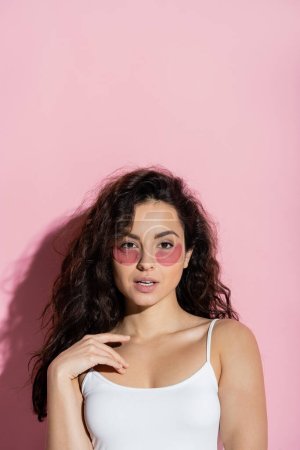 Curly brunette woman with hydrogel eye patches touching chest on pink background 